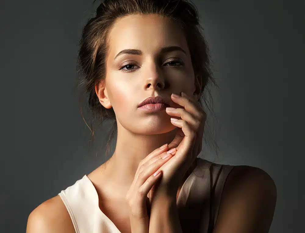 Beautiful young woman with smooth, radiant skin holds one hand up to her mouth modeling for the Schedule a Consultation section of the Xeomin service page.