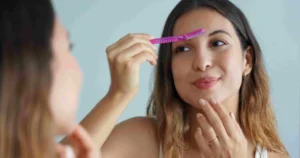 A smiling woman with wavy brown hair grooms her eyebrows with a pink razor while looking in a mirror during a dermaplaning session in Henderson, Las Vegas, NV.