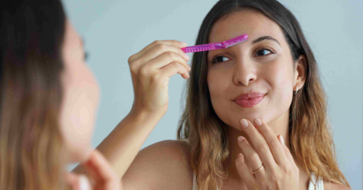 A smiling woman with wavy brown hair grooms her eyebrows with a pink razor while looking in a mirror during a dermaplaning session in Henderson, Las Vegas, NV.